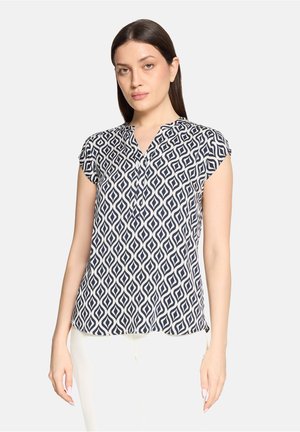 Vrouw met steil donker haar, gekleed in een kortemouwige marineblauwe en witte blouse met geometrisch patroon en een witte broek, staand tegen een effen achtergrond.