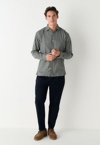 Hemd mit Knopfleiste im Karo-Muster in Grün, Marineblau und Weiß; kombiniert mit marineblauen Hosen und braunen Wildlederschuhen.