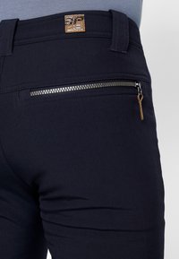 Detailní záběr na zadní kapsu námořnických kalhot s horizontálním stříbrným zipem a hnědým koženým poutkem, které jsou nošeny se zasunutou šedou košilí.