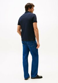 Navy poloshirt met korte mouwen, gecombineerd met blauwe denim jeans. Model staat met een lichte draai, waardoor een schone, minimalistische uitstraling zichtbaar is.