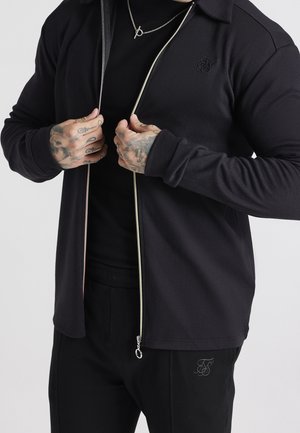 Personne aux mains tatouées fermant une veste noire sur une chemise noire, portant un collier argenté avec un fermoir à bascule.