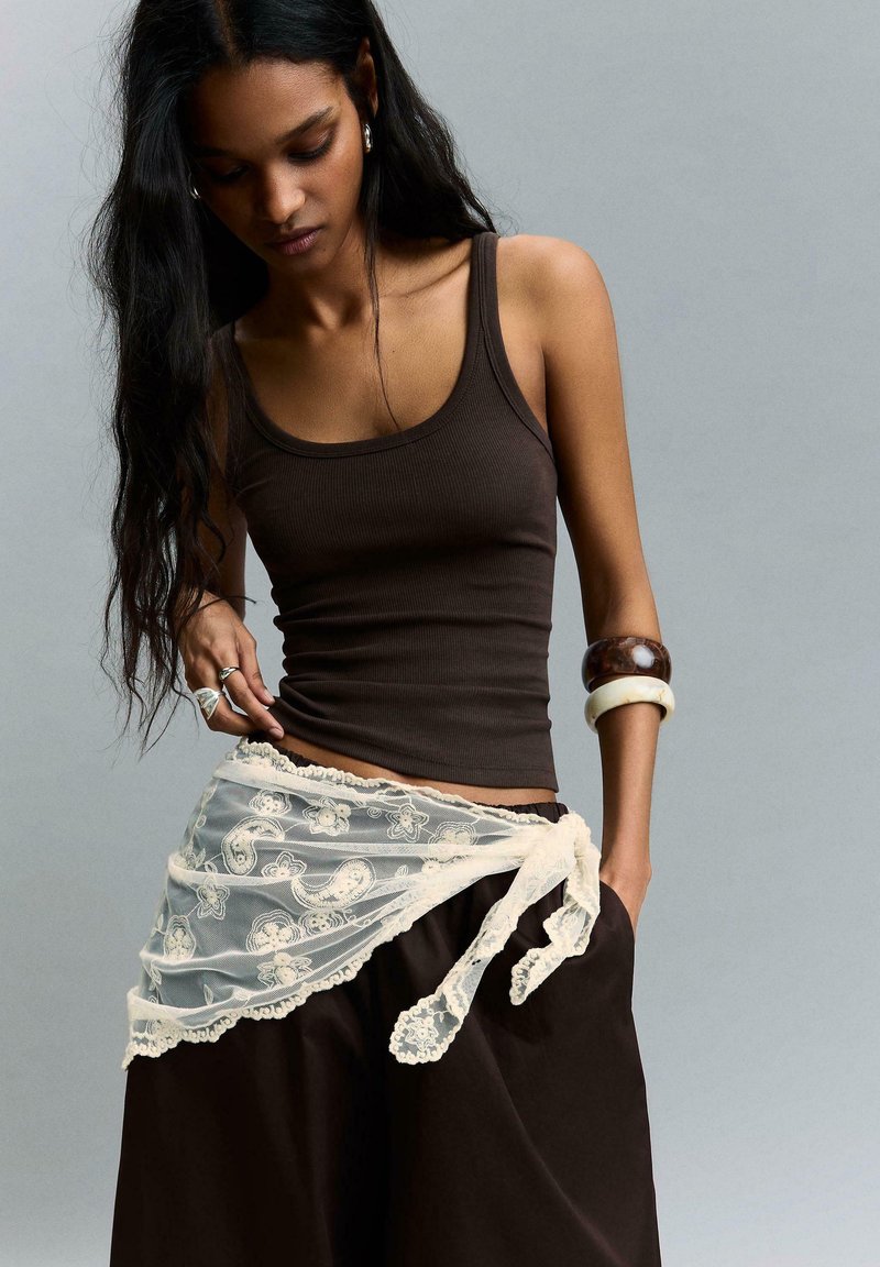 Femme aux longs cheveux foncés porte un débardeur marron ajusté, une écharpe en dentelle transparente nouée autour de la taille et de larges bracelets sur un bras.