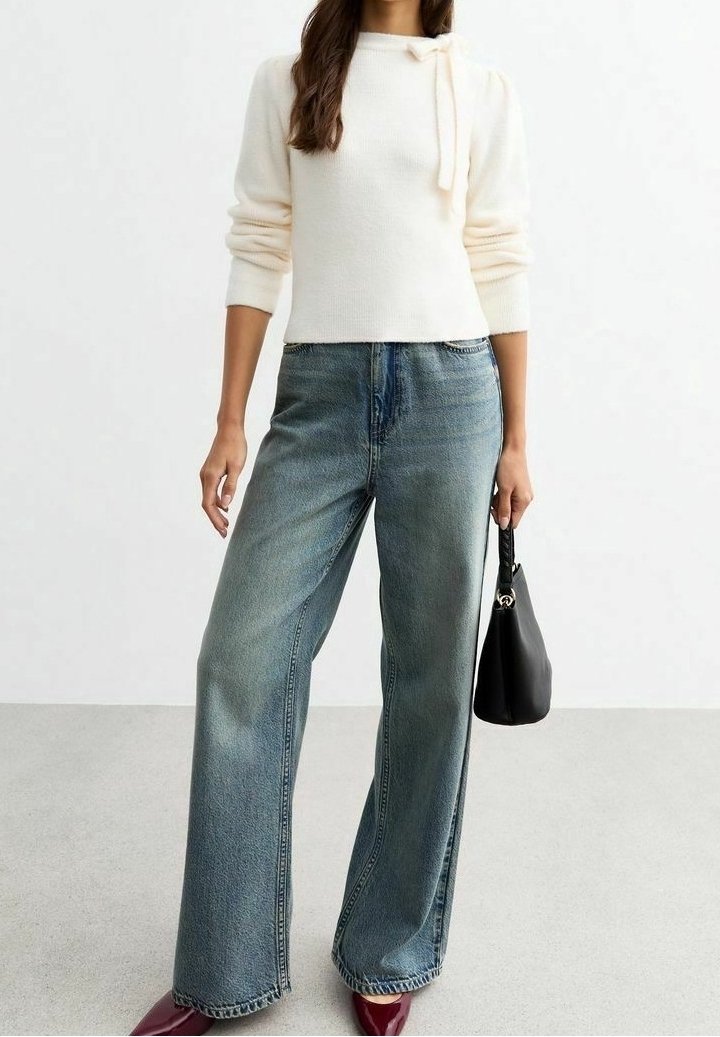 Kremowy dzianinowy sweter z wiązanym dekoltem, zestawiony z luźnymi, niebieskimi dżinsami. Dodatki to mała czarna torebka i bordowe buty.