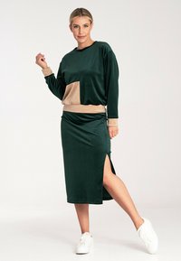 Zielona welurowa bluza z beżową kieszenią, zestawiona z dopasowaną midi spódnicą z bocznym rozcięciem. W zestawie białe sneakersy.