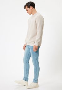 Ljust beige sweatshirt med rund hals, kombinerad med ljusblå smal passform jeans och vita sneakers. Texturerad tyg, enkel design.