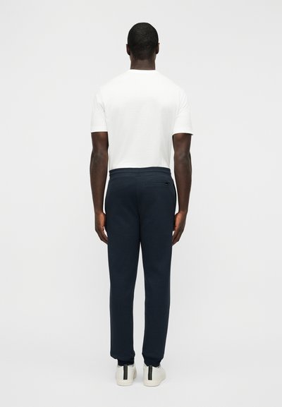 Ανδρική εμφάνιση με απλό λευκό μπλουζάκι και σκούρο μπλε φόρμα. Το παντελόνι έχει πίσω τσέπη και στενεύει στους αστραγάλους. Λευκά αθλητικά παπούτσια.