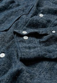 Chemise en tissu bleu foncé avec un tissage texturé, dotée de boutons en plastique blanc et d'un col rabattu. Des poches sont visibles à l'avant.