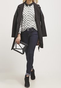 Kobieta idąca, ubrana w czarny płaszcz na kraciastą koszulę, ciemne skinny jeans, czarne botki za kostkę, trzymająca geometryczną torebkę w czarno-białe wzory.