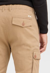 Khaki cargo bukser med knaplukning på lommer, elastisk talje og glat bomuldsstof. Bemærkelsesværdige detaljer inkluderer to knapper.
