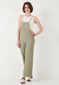 Giocattolo verde oliva, con texture, tuta con spalline regolabili e tasche laterali. Indossata sopra una canotta bianca, abbinata a sandali bianco e neri.