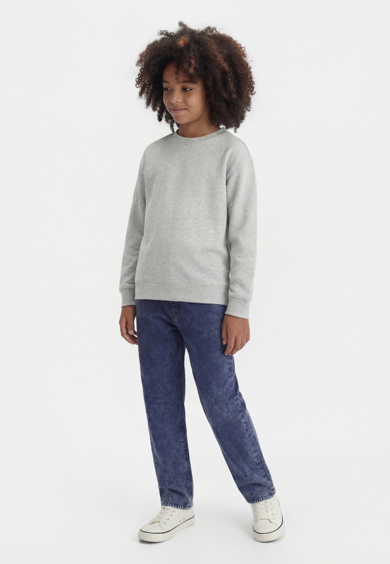 Barn med krøllete hår iført en lys grå genser, blå jeans og hvite joggesko, stående med et lett smil mot en hvit bakgrunn.