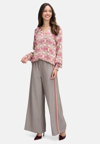 Langärmlige Bluse im rosa und grünen geometrischen Muster; kombiniert mit weit geschnittenen grauen Hosen mit Nadelstreifen und farbigen seitlichen Akzenten.