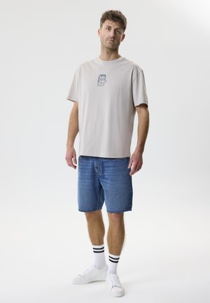 Férfi áll, bézs színű grafikás pólót, kék farmer rövidnadrágot, fekete csíkos fehér zoknit és fehér tornacipőt viselve, egyszínű háttér előtt.