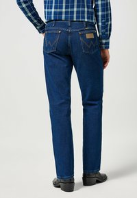 Personne portant un jean straight-leg bleu foncé avec des poches arrière et des chaussures à talons noires, vêtue d'une chemise à carreaux bleue.