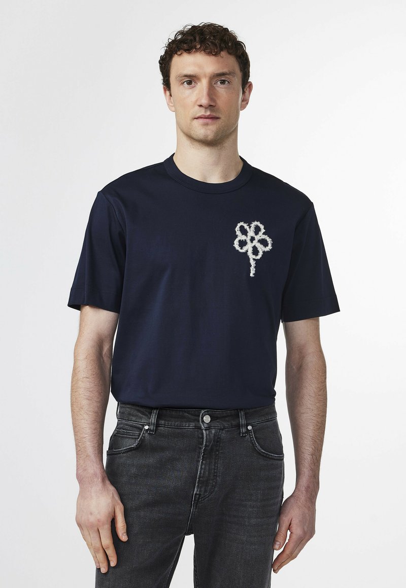Navy blue cotton t-shirt with short sleeves featuring a textured white flower design on the left chest. Worn with dark jeans.