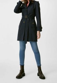 Trench-coat bleu marine avec ceinture à la taille, boutons croisés et poches inclinées. Porté avec un jean skinny bleu clair et des bottes noires épaisses.