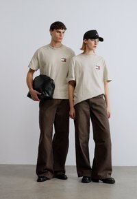 Μπεζ μπλουζάκια με ένα μικρό γραφικό στο στήθος, φαρδιά καφέ παντελόνια και μαύρο καπέλο. Ένα μοντέλο κρατάει μια μαύρη τσάντα.