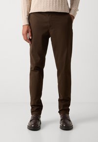 Man wearing dark brown slim-fit pants, light beige cable-knit sweater, and dark brown leather loafers, standing on plain white floor.