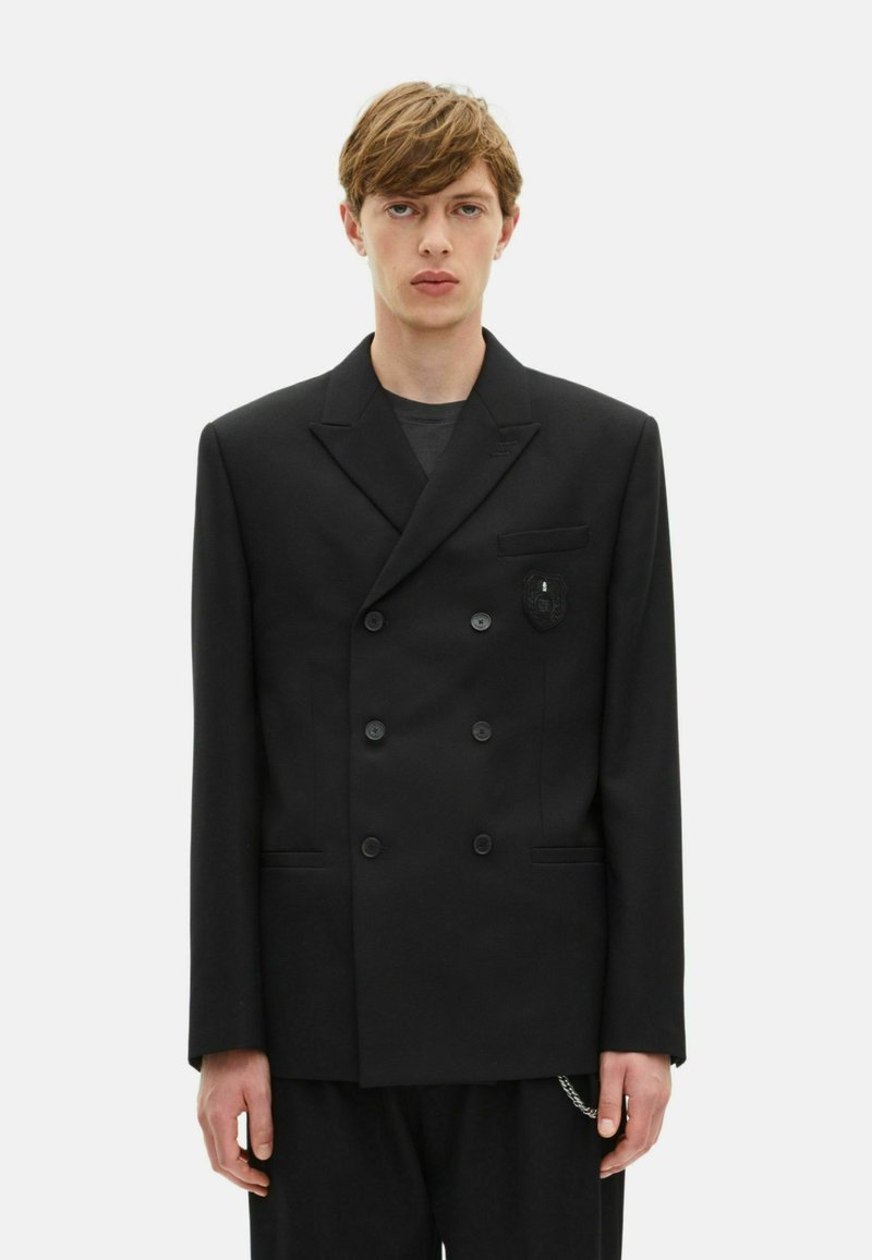 Zwarte dubbelknop blazer van gladde stof. Heeft een borstzak met een logo patch, geknipte revers en knoopdetails.