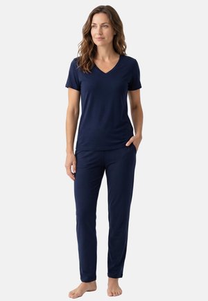 Woman standing barefoot wearing navy blue short-sleeve V-neck top and matching pants, hand in pocket, looking slightly to the side.