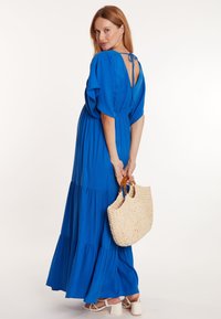 Robe longue bleue avec des manches flottantes, au design à volants, dos ouvert avec un système de fermeture à nouer. Le mannequin porte un sac en osier et des sandales à talons blocs blanches.