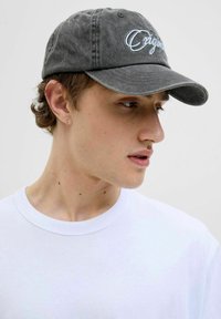 Grå bomuldsbaseballkasket med broderet "Original" tekst, buet skygge og sekspanelsdesign, båret af en person i en hvid t-shirt.