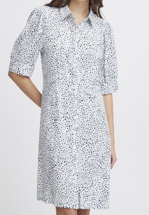 Biała sukienka z nieregularnym wzorem czarnych i niebieskich kropek, z krótkimi rękawami i kołnierzykiem, noszona przez osobę z jedną ręką zgiętą za plecami.