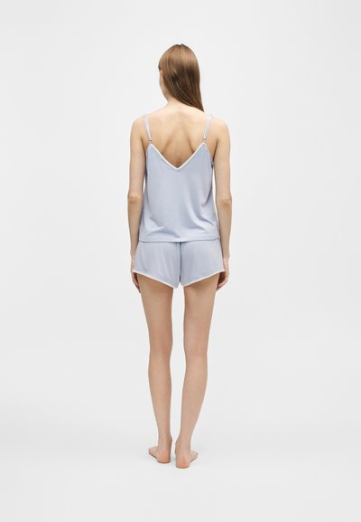 Femme debout pieds nus de dos, portant un ensemble pyjama débardeur et short bleu clair avec fines bretelles et bordure en dentelle.