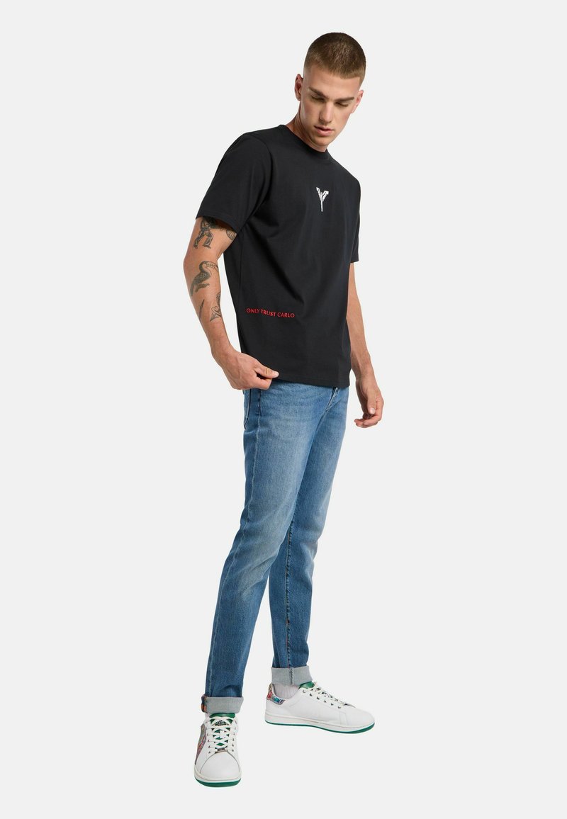 Sort bomulds t-shirt med et lille hvidt logo på brystet og rød tekst "ONLY TRUST CARLO" nær kanten, parret med lyseblå jeans og hvide sko.