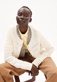 Cremfarbener, gestrickter Cardigan mit strukturiertem Muster, Holzknöpfen, übereinander getragen mit einem gelben Kragenhemd und braunen, weit geschnittenen Hosen.