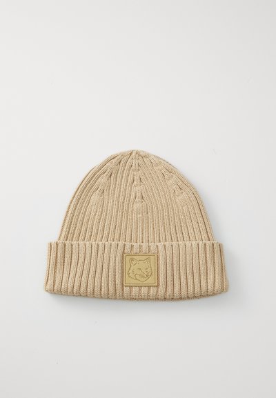 Bonnet beige en tricot avec un rebord replié et un écusson en forme de carré arborant un design de renard. Côtelé texturé sur l'ensemble du matériau.