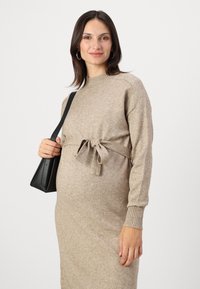 Abito lavorato a maglia beige con maniche lunghe e un fiocco in vita. La modella tiene una piccola borsa nera, caratterizzata da una texture liscia.