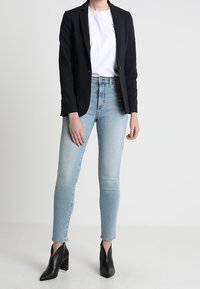 Blazer negro ajustado sobre una camiseta blanca, combinado con jeans ajustados de mezclilla azul clara y botines de tacón negros. Tejido suave y diseño ajustado.