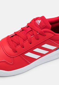 Zapatilla deportiva roja con parte superior de cuero, detalles en tejido texturado, logo de tres rayas blancas y cordones planos rojos. Suela blanca visible.