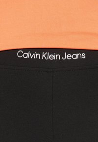 Černé legíny s elastickým materiálem a hladkou texturou, s bílým vyšitým logem "Calvin Klein Jeans" na pasu, v kombinaci s oranžovým topem.