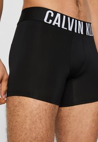 Black cotton boxers with a wide elastic waistband featuring white "CALVIN KLEIN" logo. Smooth texture, fitted design, and mid-thigh length.