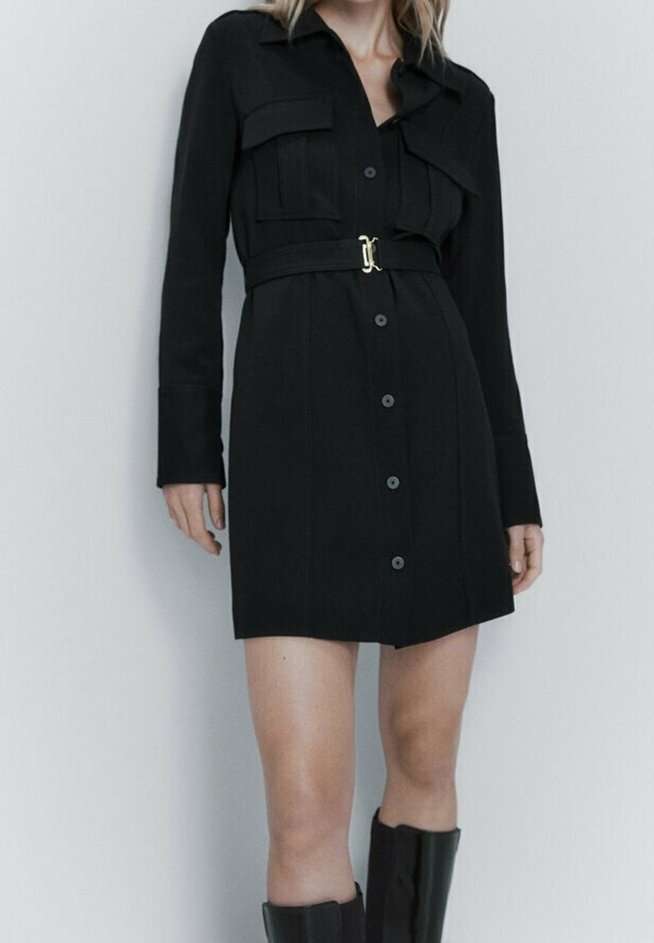 Robe chemise noire à manches longues, dotée de deux poches poitrine, d'une taille ceinturée avec une boucle dorée, et d'un devant à boutons.