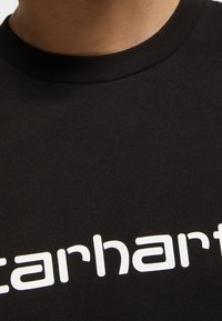 Nahaufnahme eines schwarzen Rundhalsausschnitt-T-Shirts mit weißem "Carhartt"-Aufdruck auf der Brust, getragen von einer Person mit sichtbarem Hals.
