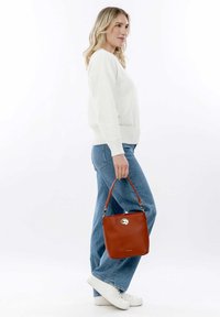 Bolso de cuero rojo con forma de cuadrado redondeado, asa corta, herrajes plateados y superficie texturizada, sostenido por una persona con atuendo casual.