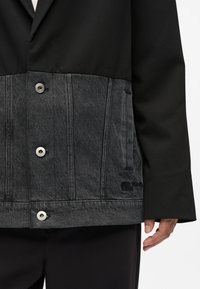 Crna jakna s donjim panelom od sive denimske tkanine. Ima metalne kopče i teksturiranu tkaninu. Istaknut logo utonut u donji deo.