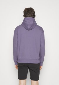 Felpa con cappuccio viola dalla texture morbida, maniche lunghe e orlo inferiore largo, abbinata a pantaloni corti neri. Vista posteriore mostrata.