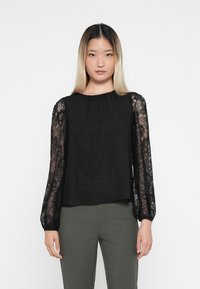 Svart topp med strukturert kropp og lange ermer i svart blonde. Rund halsutskjæring. Sammen med olivengrønne bukser. Helt hvit bakgrunn.