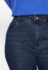 Vaqueros de cintura alta de un azul oscuro con una textura de mezclilla suave, costuras amarillas, bolsillos frontales y un cierre de botón metálico.