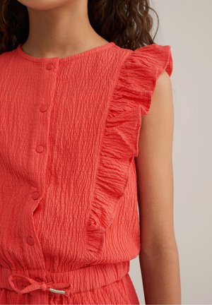 Fille portant une robe corail sans manches texturée avec des boutons recouverts de tissu, un détail à volants sur les épaules et une taille à cordon.