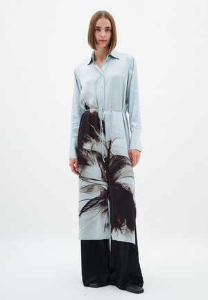 Helles blaues Satinhemdkleid mit einem dunklen Blumenmuster, langen Ärmeln, Knopfleiste und einem Taillenzug, kombiniert mit weit geschnittenen schwarzen Hosen.