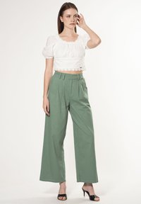 Witte, gestructureerde crop top met een gegolfde zoom en pofmouwen, gecombineerd met hooggewaaiste, wijde groene pantalons en zwarte open teen hakken.