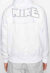 Bílá větrovka s kapucí, s velkým vyšívaným logem "NIKE" na zádech, elastickými manžetami a volným střihem.