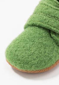 Zielone filcowe kapcie z teksturowaną górą i gumową podeszwą. Charakteryzują się miękkim wykończeniem oraz widocznymi detalami w postaci szwów.