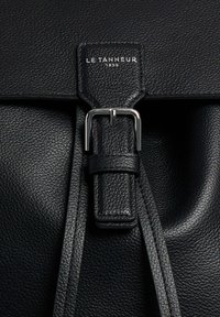 Primer plano de un bolso de cuero negro que presenta una hebilla de metal plateado y una superficie texturizada. La marca "LE TANNEUR 1898" está grabada en el solapa.