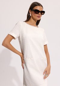 Kobieta ubrana w białą teksturowaną sukienkę z krótkim rękawem i perłowym wykończeniem, duże kolczyki, ogromne czarne okulary przeciwsłoneczne oraz wyróżniający się pierścionek, pozująca z ręką na biodrze.